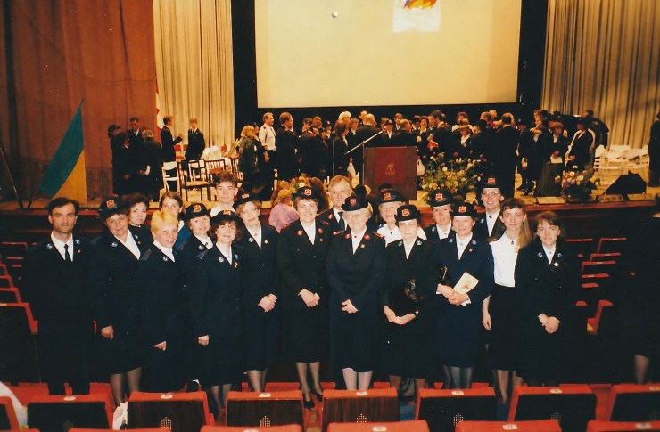 Gruppebillede af kvinder og mænd i uniformer med medaljer, der står foran en scene ved en festlig begivenhed.